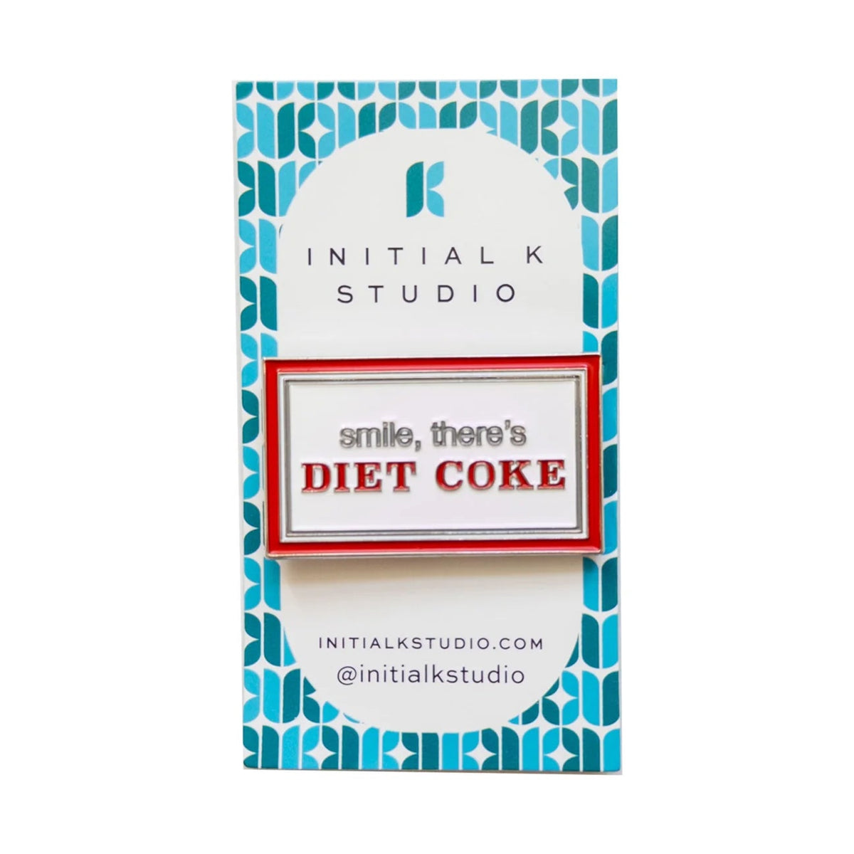 An enamel pin reading 'smile, there's DIET COKE' in red and gray lettering, mounted on a white card with a blue geometric pattern. The card is branded 'INITIAL K STUDIO'.