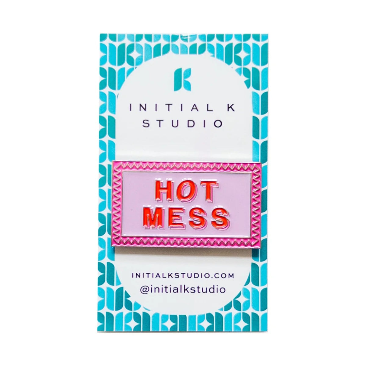 An enamel pin spelling 'HOT MESS' in red letters on a pink background with a pink zigzag border, mounted on a white and teal patterned display card from Initial K Studio.