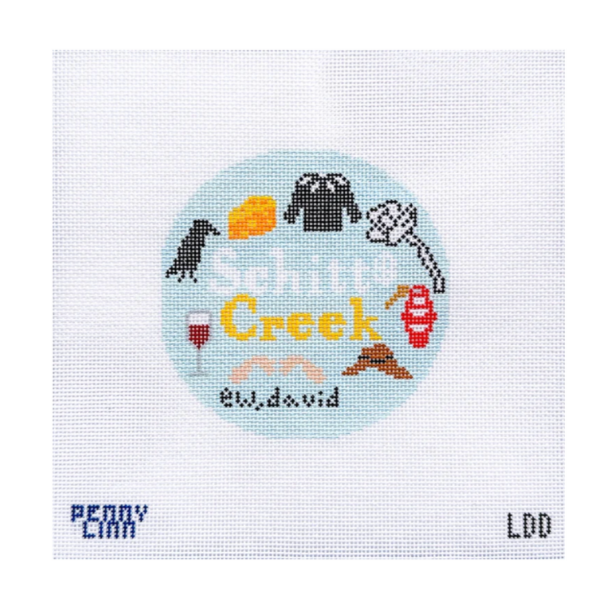 A square cross-stitch design on white Aida fabric, featuring a circular light blue patch with the words 'Snipitit Creek' in yellow, surrounded by small embroidered icons in black, red, orange, brown, and white. The bottom of the fabric shows the text 'PERRY CANN' on the left and 'LDD' on the right.