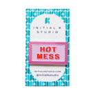An enamel pin spelling 'HOT MESS' in red letters on a pink background with a pink zigzag border, mounted on a white and teal patterned display card from Initial K Studio.