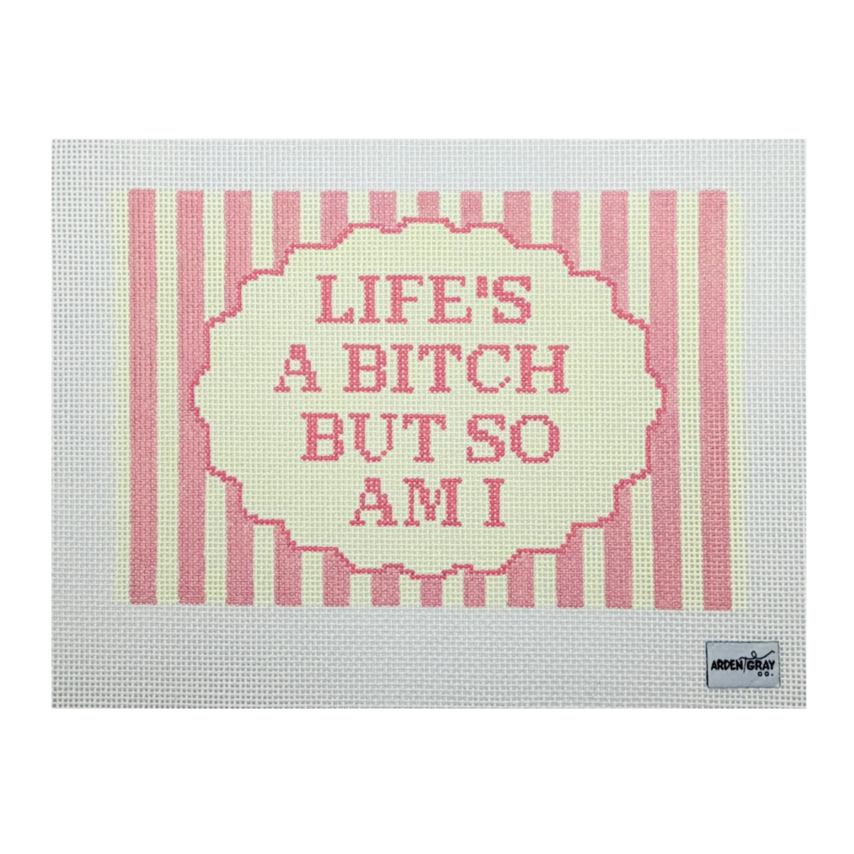 A cross-stitch canvas featuring the text 'LIFE'S A BITCH BUT SO AM I' in pink within a scalloped cream border, set against pink and cream vertical stripes on a white Aida cloth. The 'another' brand logo is visible in the bottom right corner.