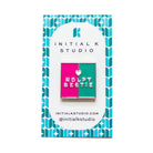 A square enamel pin by Initial K Studio featuring a split design of hot pink and teal with the text 'NDLP BESTIE' and a small heart, mounted on a branded white backing card with a blue geometric border.