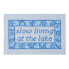 A square needlepoint canvas featuring the phrase 'slow living at the lake' in blue lettering, surrounded by a decorative blue border on a white grid background.