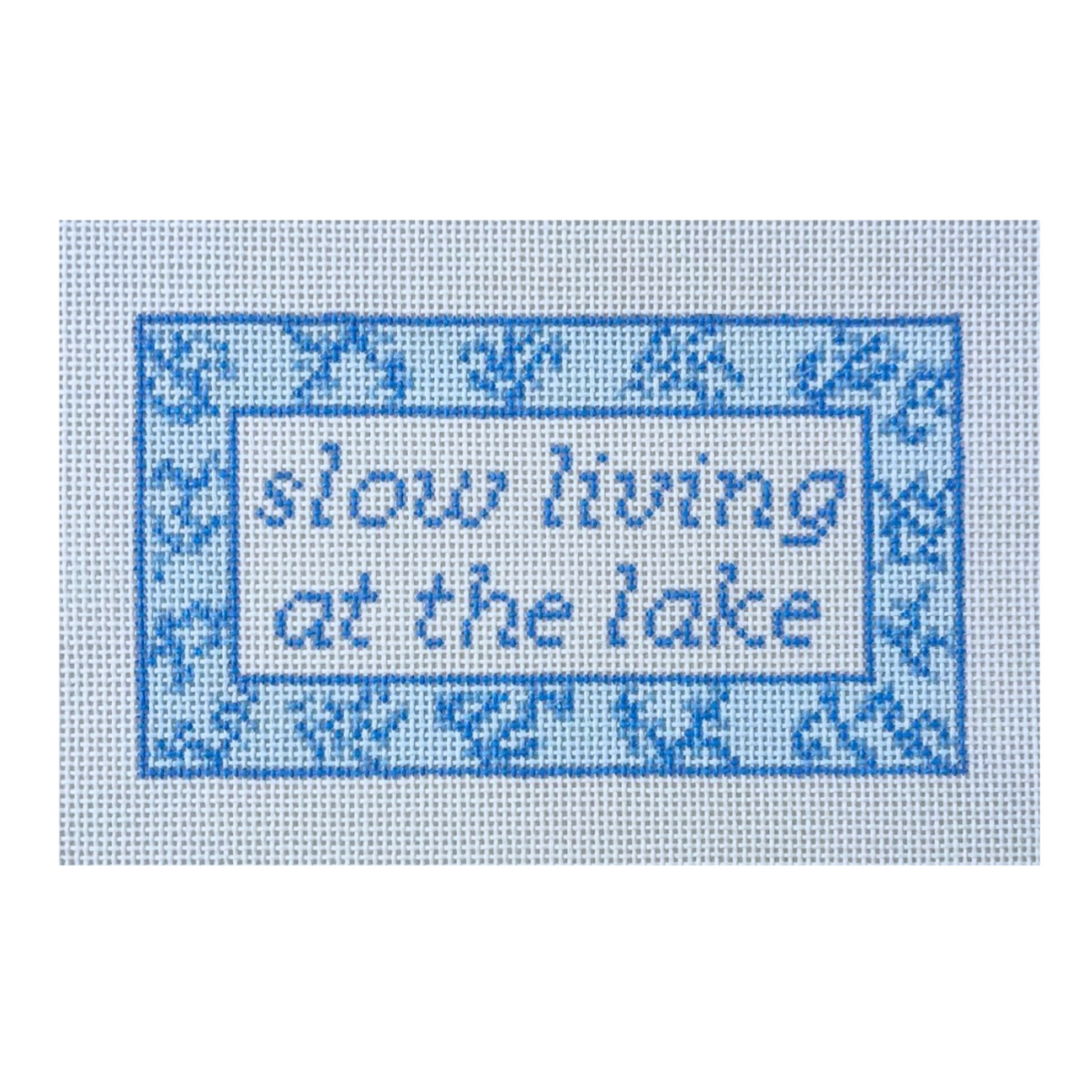 A square needlepoint canvas featuring the phrase 'slow living at the lake' in blue lettering, surrounded by a decorative blue border on a white grid background.