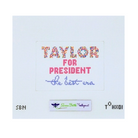 A cross-stitch style design on a white fabric background reading 'TAYLOR FOR PRESIDENT the best era' in multicolored, pink, and blue lettering, with a small Green Belt United States logo below.