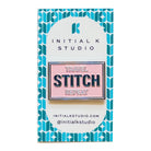 A rectangular enamel pin on a branded backing card. The pin has a pink background with navy and white text reading 'TRAVEL STITCH REPEAT.