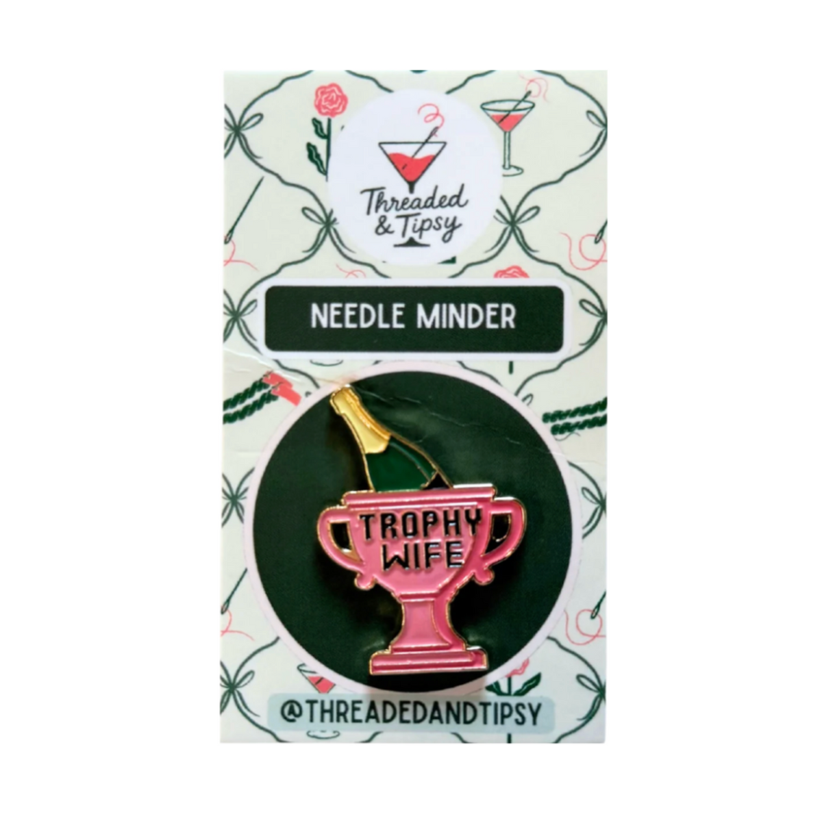 A pink enamel pin shaped like a trophy cup with the words 'TROPHY WIFE' on it, attached to a branded backing card. The card reads 'Threaded & Tipsy' and 'Needle Minder,' with decorative green vine illustrations on a white background.