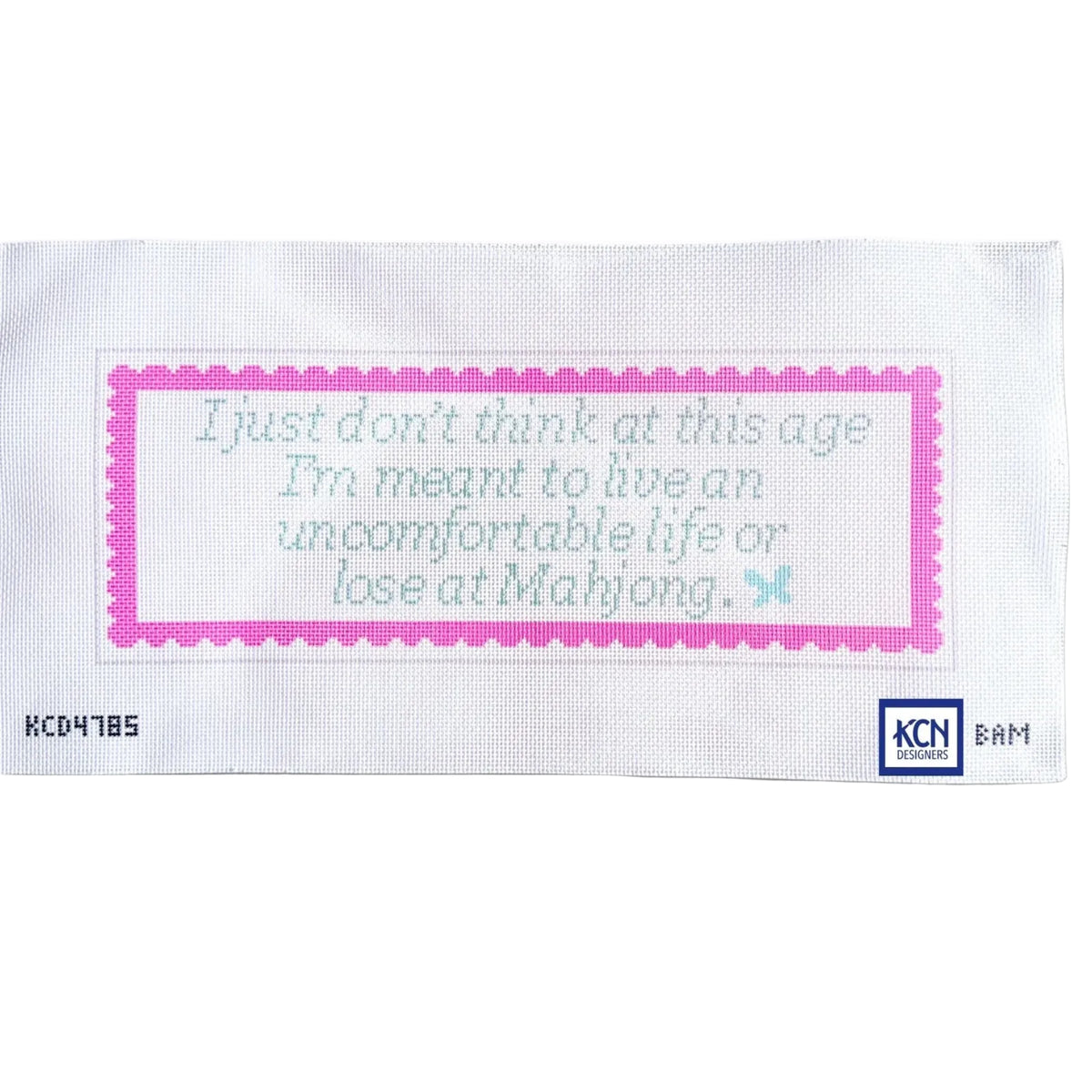 A white rectangular cross-stitch sampler card featuring a pink scalloped border and light blue embroidered text that reads, 'I just don't think at this age I'm meant to live an uncomfortable life or lose at Mahjong.' The card has a small KCN Mahjong logo in the bottom right corner and a serial number on the bottom left.