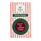 A circular enamel pin with the text 'This is my stabbing hobby' in white on a dark red center, surrounded by a dark green ring, mounted on a pink and white striped backing card labeled 'Threaded & Tipsy Needle Minder'.